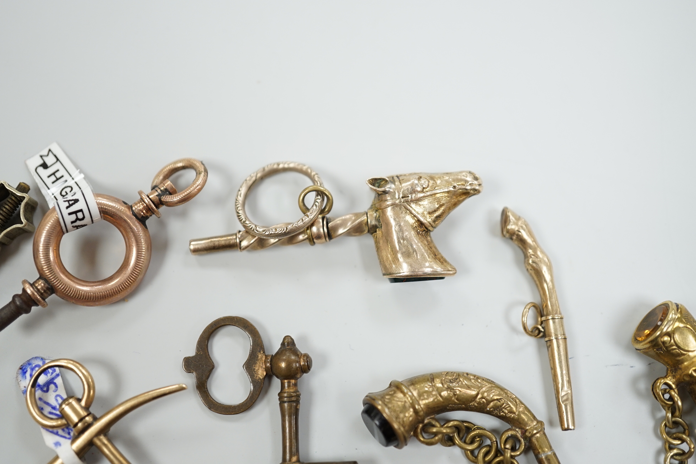 Eighteen assorted mainly 19th century base or yellow metal overlaid novelty watch keys, including coral and bloodstone set harp, pistol, rifle, cornucopia, bellows, horses hoof and trade insignia, largest 55mm.